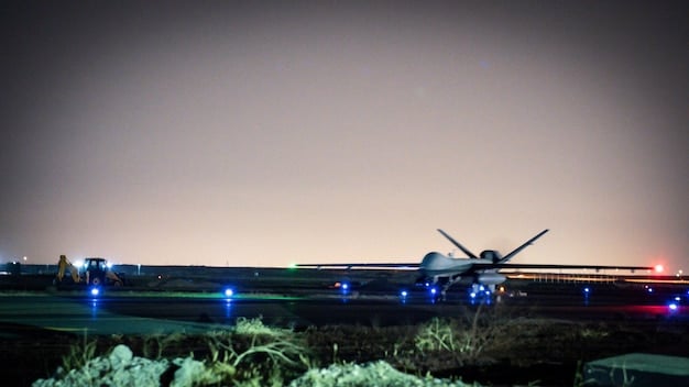 A digital rendering of a futuristic military drone, showcasing advanced sensors and weaponry. The background is a blurred cityscape at night, suggesting urban warfare scenarios.
